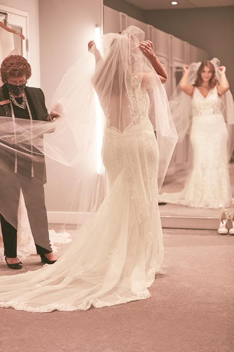 bride wearing a veil and wedding dress looking into a mirror as store associate holds veil