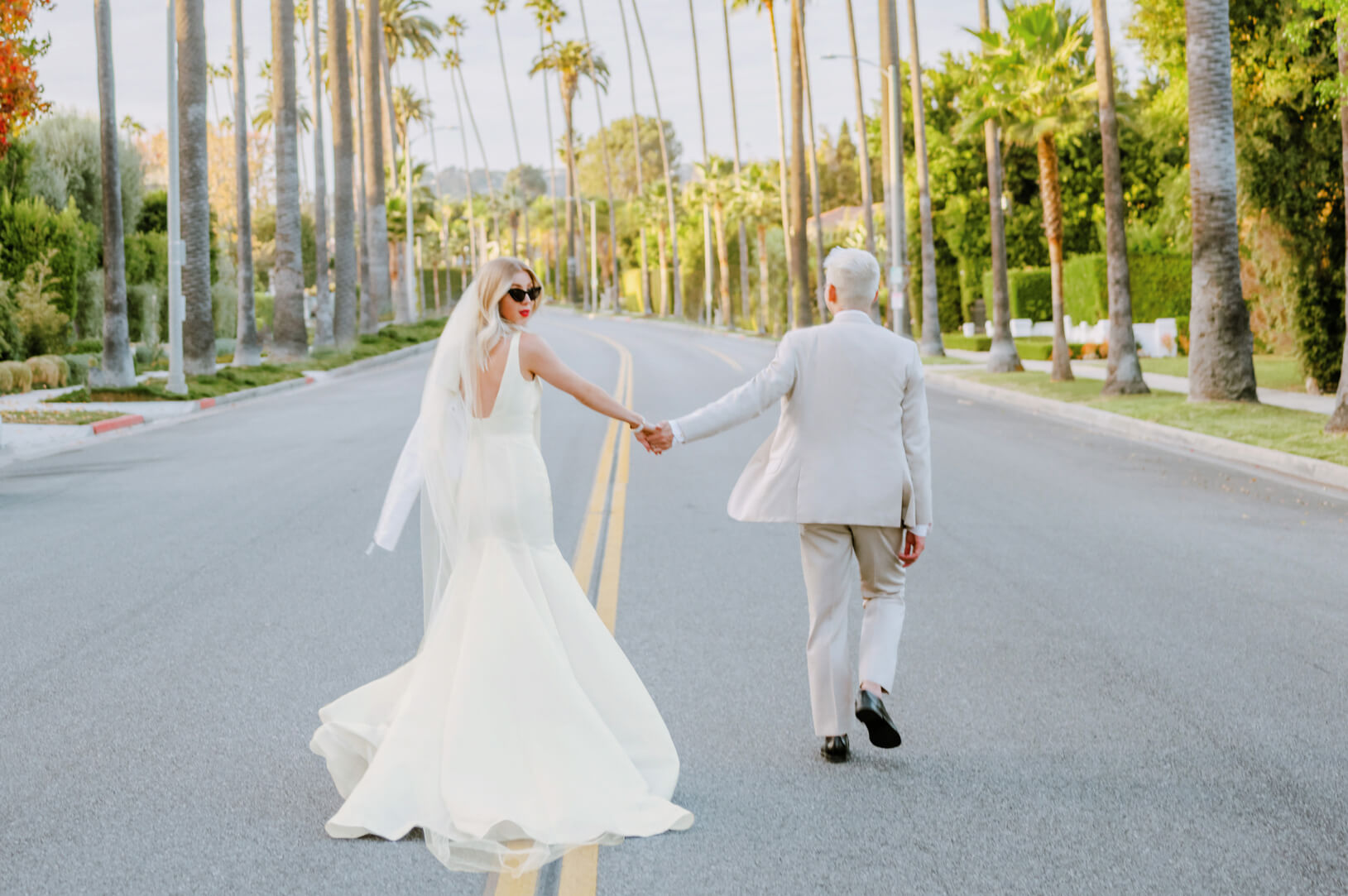 bride wearing wedding dress and sunglasses holding hands with groom and walking in street