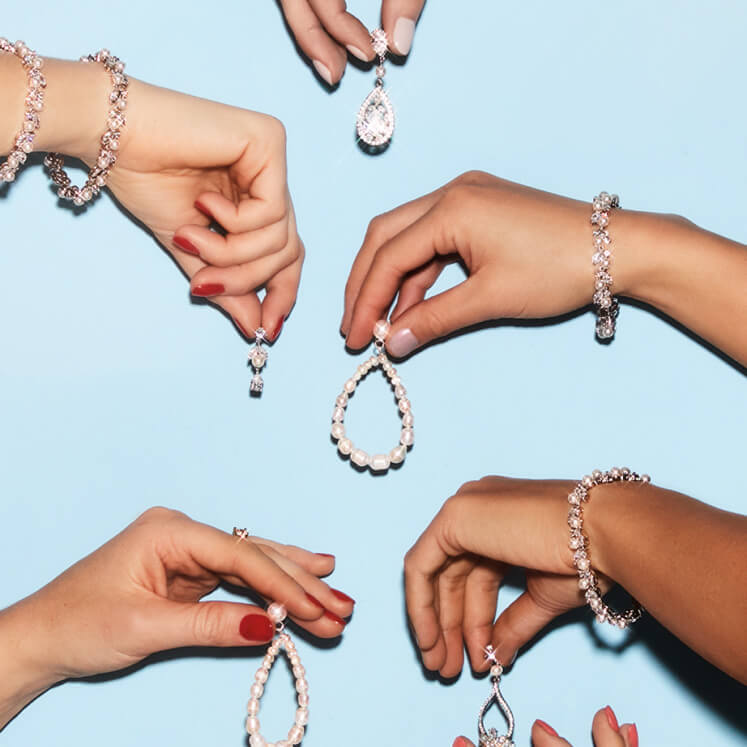 several pairs of hands wearing diamond bracelets holding diamond and pearl earrings