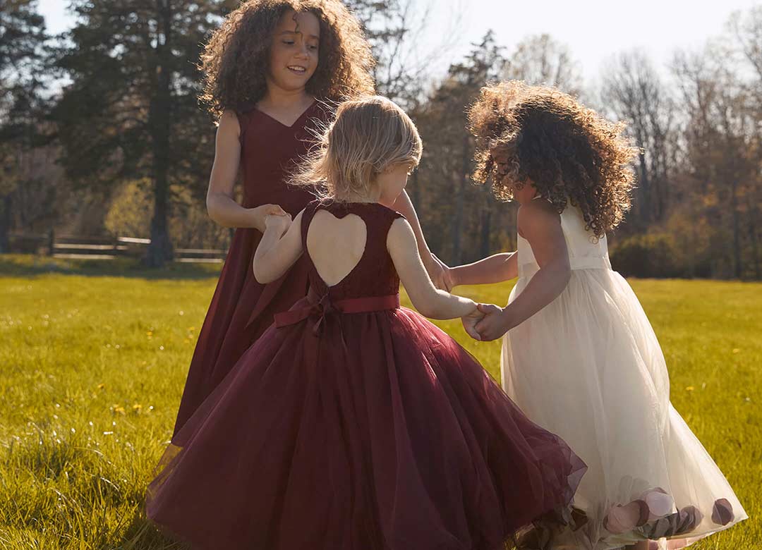3 flower girls in wine and white dresses holding hands in a field