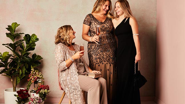 woman in pantsuit sitting next to 2 women in taupe and black dresses standing in a room