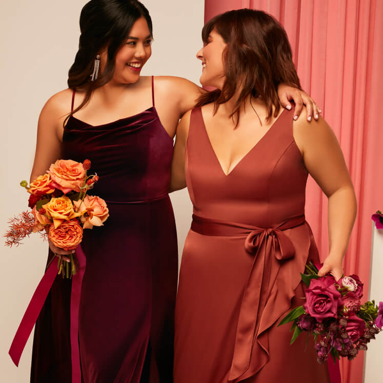 two brides in maroon and red dresses with arms around each other and holding flower bouquets