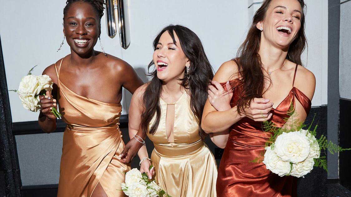 three bridesmaids in different color dresses walking with arms locked smiling and holding flower bouquets