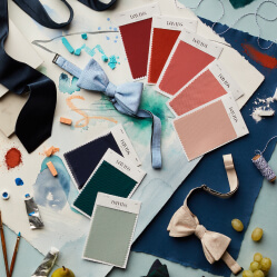 multiple fabric swatches on a table next to bow ties, neckties, paint, and paint brushes