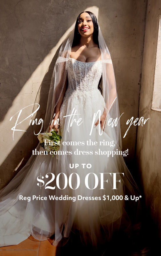 woman wearing a wedding dress leaning against a wall holding a bouquet promoting ring in the new year first comes the ring, then comes the dress shopping! up to $200 off reg price wedding dresses $1,000 & up*